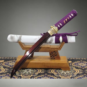 May include: A short sword with a dark red blade and a purple wrapped handle featuring gold-coloured accents. The sword rests on a wooden stand behind a white scabbard with a purple cord tie. The handle has a diamond-patterned wrap over white.
