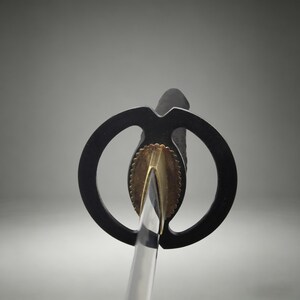 May include: A Japanese sword featuring a circular black tsuba handguard, a metallic gold-toned habaki collar with a scalloped edge, and a portion of the polished steel blade visible below the guard, extending from a black textured handle grip.