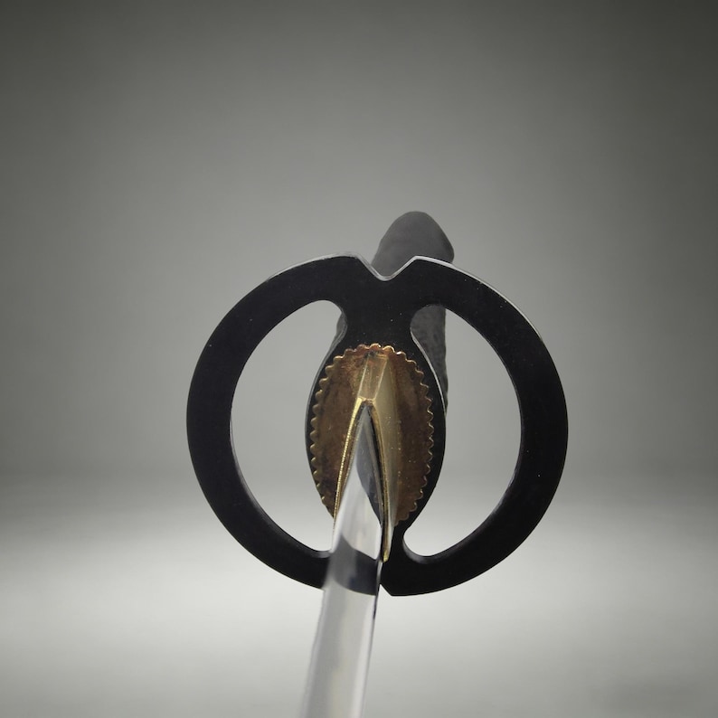 May include: A Japanese sword featuring a circular black tsuba handguard, a metallic gold-toned habaki collar with a scalloped edge, and a portion of the polished steel blade visible below the guard, extending from a black textured handle grip.