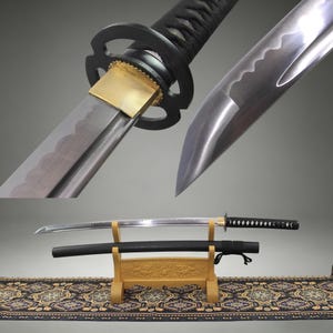 Può includere: Una spada katana con lama in acciaio lucido caratterizzata da una linea di tempra ondulata visibile, un collare in ottone e una guardia circolare nera. L'impugnatura è avvolta da un cordino nero. Un fodero nero abbinato poggia su un supporto in legno chiaro.