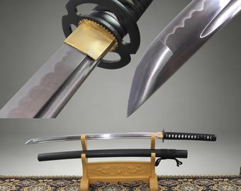 Puede incluir: Una espada katana con hoja de acero pulido que presenta una línea de temple ondulada visible, una virola de latón y una guarda circular negra. El mango está envuelto en cordón negro. Una vaina negra a juego descansa sobre un soporte de madera clara.