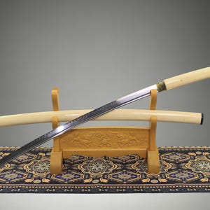 May include: A katana sword with a long, curved steel blade featuring a visible wavy temper line, a cream-colored handle, and a matching cream-colored scabbard. The sword rests on a light-colored wooden stand with carved decorative details.