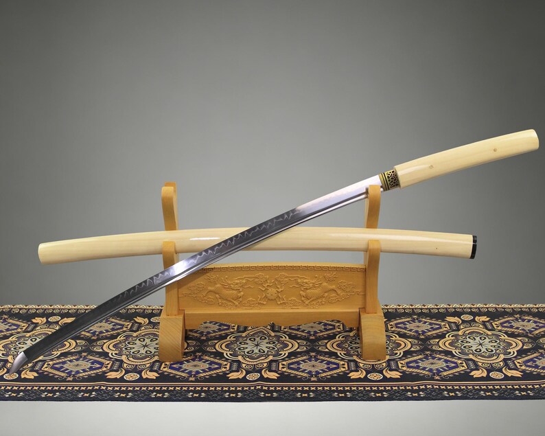 May include: A katana sword with a long, curved steel blade featuring a visible wavy temper line, a cream-colored handle, and a matching cream-colored scabbard. The sword rests on a light-colored wooden stand with carved decorative details.
