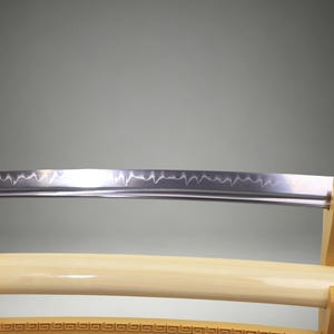 May include: A Japanese katana sword with a polished steel blade featuring a visible wavy hamon line. The sword is resting on a light-colored wooden stand, with a matching cream-colored scabbard positioned horizontally below the blade.