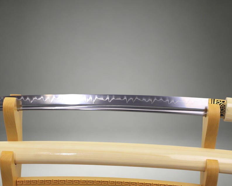 May include: A Japanese katana sword with a polished steel blade featuring a visible wavy hamon line. The sword is resting on a light-colored wooden stand, with a matching cream-colored scabbard positioned horizontally below the blade.
