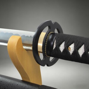 May include: A katana sword featuring a silver steel blade with a visible wavy temper line, a circular black metal tsuba guard, and a handle wrapped in black cord over white ray skin. The sword rests horizontally on a light-colored wooden stand.