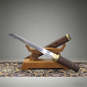 May include: A Japanese-style sword with a polished steel blade, a dark wood handle, and a matching dark wood scabbard. The sword rests on a light-colored wooden display stand featuring intricate carved details, showcasing a sharp, curved edge.