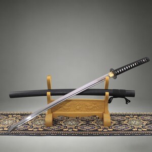 May include: A katana sword with a silver-toned blade, a black wrapped handle, and a circular guard. The sword rests on a light-colored wooden stand alongside a black scabbard. The stand features carved details and sits on a patterned fabric runner.