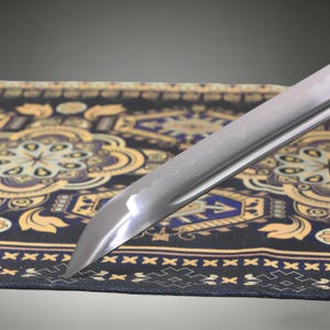 May include: A sharp, curved metal sword blade featuring a visible wavy temper line along the edge. The polished steel surface reflects light, showing a distinct point and a tapered design that extends diagonally across the frame.