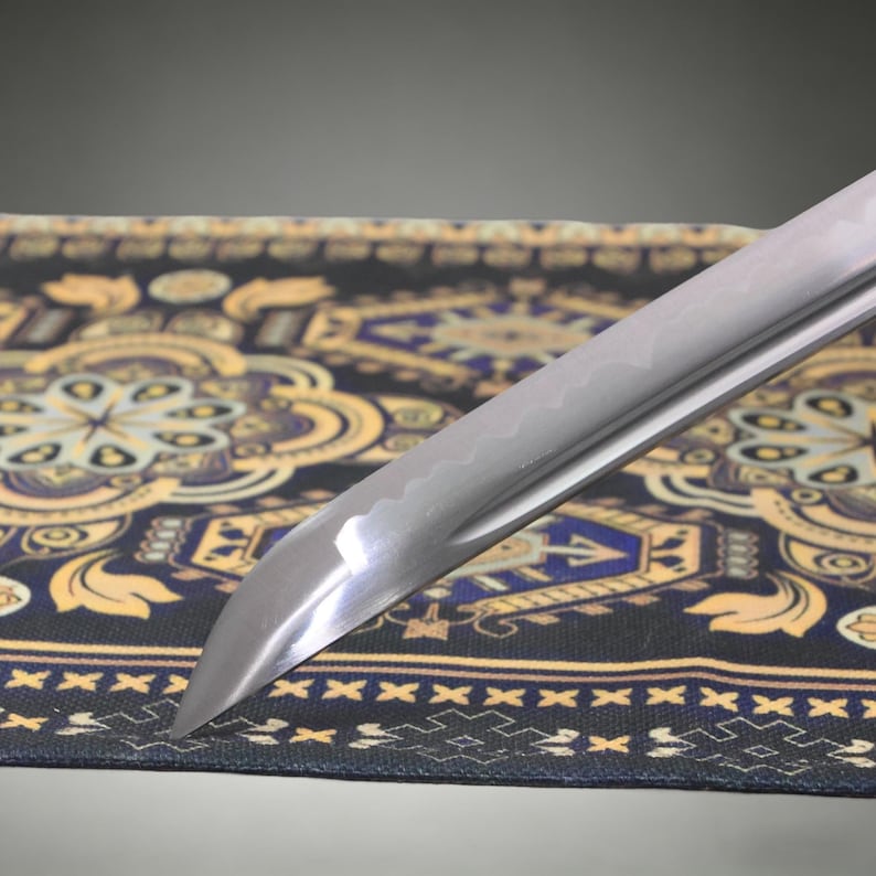 May include: A sharp, curved metal sword blade featuring a visible wavy temper line along the edge. The polished steel surface reflects light, showing a distinct point and a tapered design that extends diagonally across the frame.