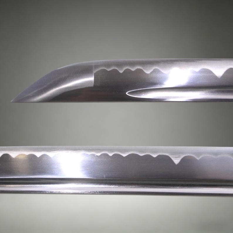 May include: A close-up view of a polished steel katana blade featuring a distinct wavy hamon line along the cutting edge. The blade includes a visible groove, known as a bo-hi, running along the side, with a sharp, curved tip at the end.