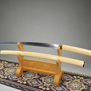 May include: A katana sword with a curved steel blade and a smooth, cream-colored handle, resting on a wooden stand. The matching cream-colored scabbard lies on the stand below the blade. The stand features intricate carvings of animals.