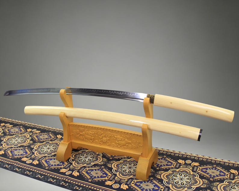 May include: A katana sword with a curved steel blade and a smooth, cream-colored handle, resting on a wooden stand. The matching cream-colored scabbard lies on the stand below the blade. The stand features intricate carvings of animals.