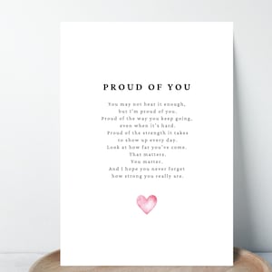 Könnte beinhalten: Eine weiße Karte mit dem Text "PROUD OF YOU" und einem inspirierenden Gedicht. Am unteren Rand der Karte befindet sich ein kleines, aquarellartiges rosa Herz. Die Karte liegt auf einem Holztablett.