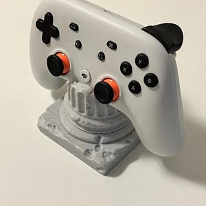 Può includere: Un controller di gioco bianco con pulsanti neri e dettagli arancioni, appoggiato su un supporto decorativo grigio. Il controller ha un pad direzionale a forma di croce e vari pulsanti funzione. Il supporto è progettato per assomigliare a una colonna classica.
