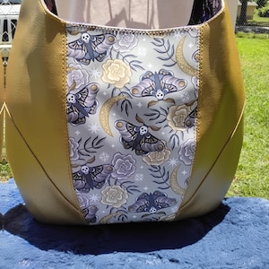 以下が含まれることがあります： マスタードイエローのトートバッグ。中央のパネルには、グレー、紫、クリーム色の蛾と花の模様が描かれています。バッグは丸みを帯びた形で、内側は濃い色です。「SCARING IS CARING」の文字が上部に表示されています。