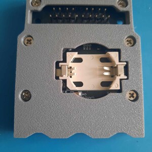 Puede incluir: Una caja de plástico gris con una placa de circuito dentro. La caja tiene una ranura para una batería y una fila de pines en la parte superior. La caja está diseñada para contener un pequeño dispositivo electrónico.