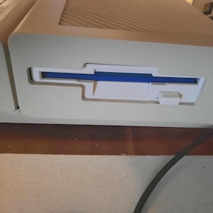 Könnte beinhalten: Nahaufnahme eines beigefarbenen Vintage-Computers mit einem Diskettenlaufwerk. Das Laufwerk hat einen blauen Schlitz und eine weiße Auswurftaste. Der Computer steht auf einer braunen Oberfläche.