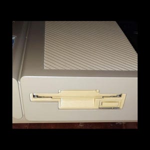 May include: Close-up of a beige computer with a floppy disk drive. The drive has a rectangular slot and a beige eject button. The top of the computer has a patterned design.