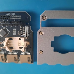 May include: A black circuit board with a white plastic component and three gold connectors. The board has the text "JLCPCB" printed on it. A gray plastic case with a cutout for the circuit board.