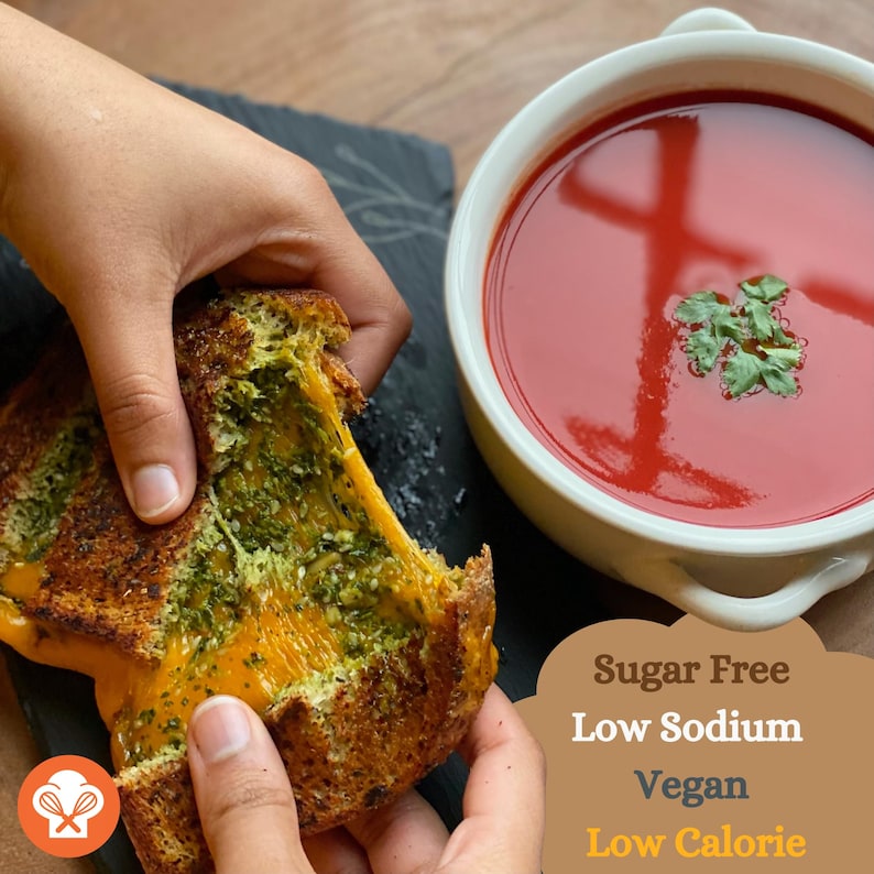 May include: A close-up of a grilled cheese sandwich with pesto and melted cheese. The sandwich is being held in two hands and a bite has been taken out of it. The sandwich is next to a bowl of red tomato soup. The text "Sugar Free Low Sodium Vegan Low Calorie" is in the bottom right corner of the image.