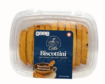 May include: A clear plastic container filled with sliced Cotto Biscottini Toasted Almond cookies. The dark blue label features a volcano illustration, nutritional facts, and the text Cotto Biscottini, High Protein, Low Sugar, Non-GMO, 5.6oz.