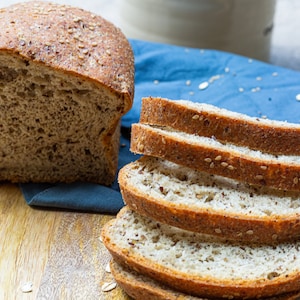 May include: A loaf of sliced multigrain bread with sesame seeds on a wooden cutting board. The bread is next to a blue cloth napkin.