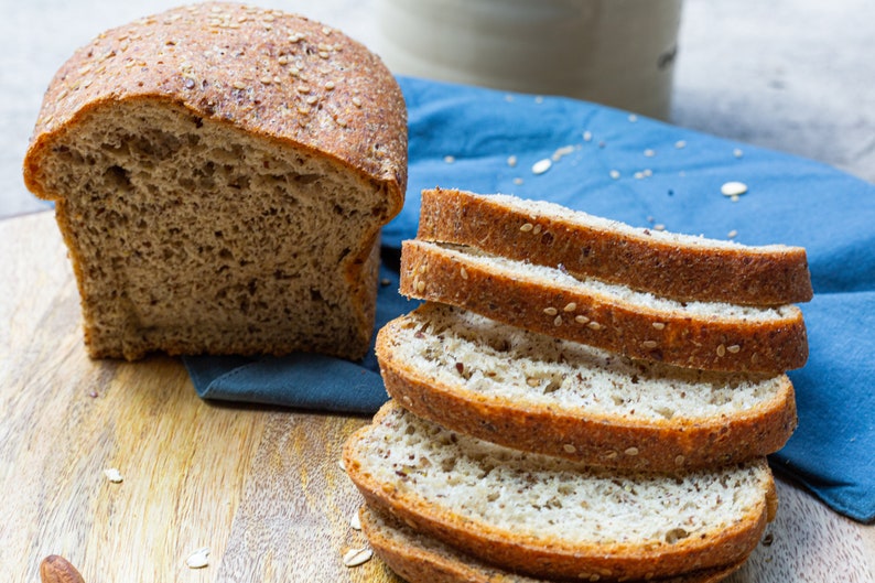 May include: A loaf of sliced multigrain bread with sesame seeds on a wooden cutting board. The bread is next to a blue cloth napkin.