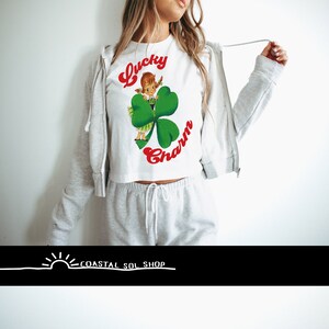 May include: White cropped t-shirt with a graphic of a cartoon girl holding a four-leaf clover. The text "Lucky Charm" is written in red letters on the shirt.