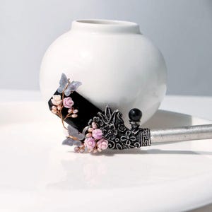 May include: A decorative hair accessory featuring a black rectangular centerpiece adorned with pink flowers, gray butterflies, and a silver floral design. The accessory has a silver handle and a black sphere at the end.
