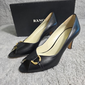 Può includere: Un paio di décolleté peep-toe in pelle nera con fibbie dorate. I tacchi a spillo sono esposti davanti a una scatola nera con la scritta "RANC" visibile. Le scarpe hanno un design classico ed elegante.
