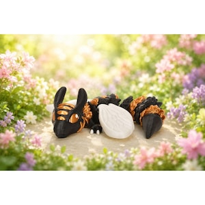 May include: A black, orange, and gold dragon figurine with large, expressive eyes and a white wing. The dragon is lying down amidst a bed of colorful flowers. The dragon's body is segmented, with a detailed texture.