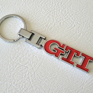 May include: A silver metal keyring featuring the red letters "GTI". The keyring has a silver ring and a rectangular clasp. The letters are outlined in silver.