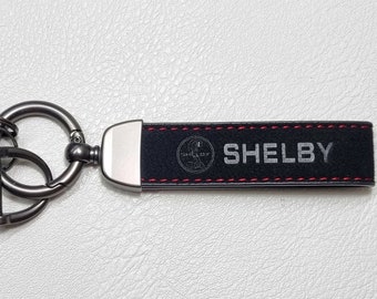 May include: Black leather keychain with the word "SHELBY" in silver lettering. The keychain has a silver metal clasp and a red stitched border. The Shelby logo is also visible.