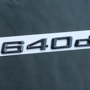 Pode incluir: Emblema preto 640d sobre fundo branco. O design tridimensional é brilhante, provavelmente para um veículo. Os números e a letra são em negrito, sem serifa, sobre uma superfície texturizada cinza escuro.