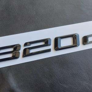 Op de afbeelding: Zwart, glanzend "320d" embleem, waarschijnlijk voor een voertuig, wordt getoond op een witte streep tegen een getextureerde grijze achtergrond. De cijfers en letter zijn in een vetgedrukte, schreefloze lettertype, wat een moderne uitstraling suggereert.