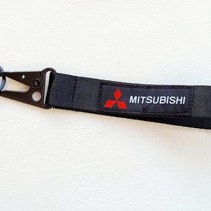 Könnte beinhalten: Schwarzes Schlüsselband mit schwarzem Metallverschluss und Schlüsselring. Der Riemen zeigt das Mitsubishi-Logo in Rot und den Markennamen in Weiß. Der Schlüsselanhänger ist für den Automobilbereich konzipiert.