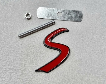 May include: Red metal 'S' emblem with a black outline. The emblem comes with a silver mounting plate, screw, and nut.