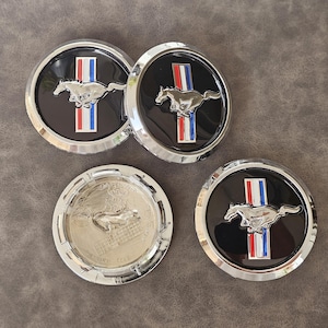 Puede incluir: Cuatro tapacubos de coche negros y plateados con acabado cromado. Cada tapacubos presenta un logotipo de caballo plateado corriendo con un diseño de rayas rojas, blancas y azules.