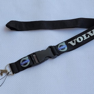 May include: Black lanyard featuring the word "VOLVO" in silver. The lanyard has a black plastic buckle and a metal clasp. The Volvo logo, in blue and silver, is printed on the lanyard.