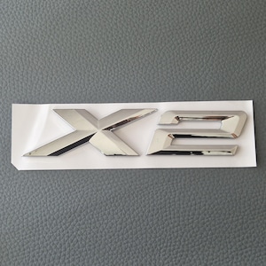 Peut inclure: Un emblème chromé "X2" sur un support blanc. Les lettres, dans une police moderne et angulaire, reflètent la lumière. L'arrière-plan est une surface grise texturée. L'emblème est probablement pour un véhicule.
