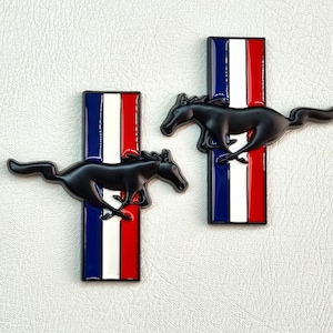 Pode incluir: Dois emblemas Mustang de metal preto com um design de cavalo correndo. Cada emblema tem um fundo de listras vermelhas, brancas e azuis.