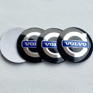 May include: Four black and silver Volvo emblem stickers with the Volvo logo in white and blue. The stickers are round and have a glossy finish. One sticker has its backing peeled off, revealing a white adhesive surface.