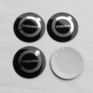 May include: Four black and silver Volvo wheel centre caps. Each cap features the Volvo logo and text in silver against a black background. One adhesive sticker is also included.