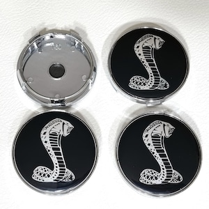 Puede incluir: Cuatro tapacubos de coche negros y plateados con un diseño de cobra plateado. La cobra está delineada en plata y tiene un fondo negro.