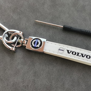 Puede incluir: Llavero de cuero blanco con un anillo de llave de metal plateado y un logotipo de Volvo de metal plateado. El llavero tiene la palabra "VOLVO" impresa en negro.