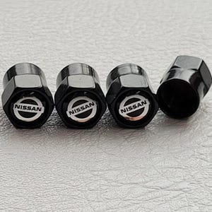 Puede incluir: Juego de cuatro tapones negros para válvulas de neumáticos. Cada tapón, de forma hexagonal, presenta el logotipo de Nissan en blanco sobre fondo negro. Un tapón está desenroscado.