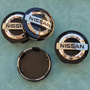 Puede incluir: Cuatro tapacubos negros para coche con un borde plateado y la palabra "NISSAN" en letras plateadas.