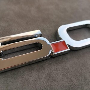 May include: Chrome-plated metal emblem with the number "5.0" in raised lettering. The "0" has a red rectangular insert.