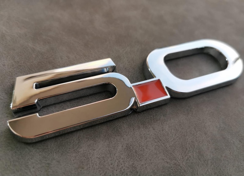 May include: Chrome-plated metal emblem with the number "5.0" in raised lettering. The "0" has a red rectangular insert.